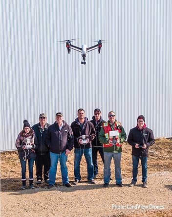 Seven people lined in with for a photo at a farm - most holding a drone controller - with a drone in the flying in the foreground