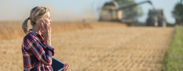 A farmer discusses the day's progress on her tablet while machinery works diligently in the background. The fields are golden and ripe, reflecting a successful harvest season under the sun's warm light.