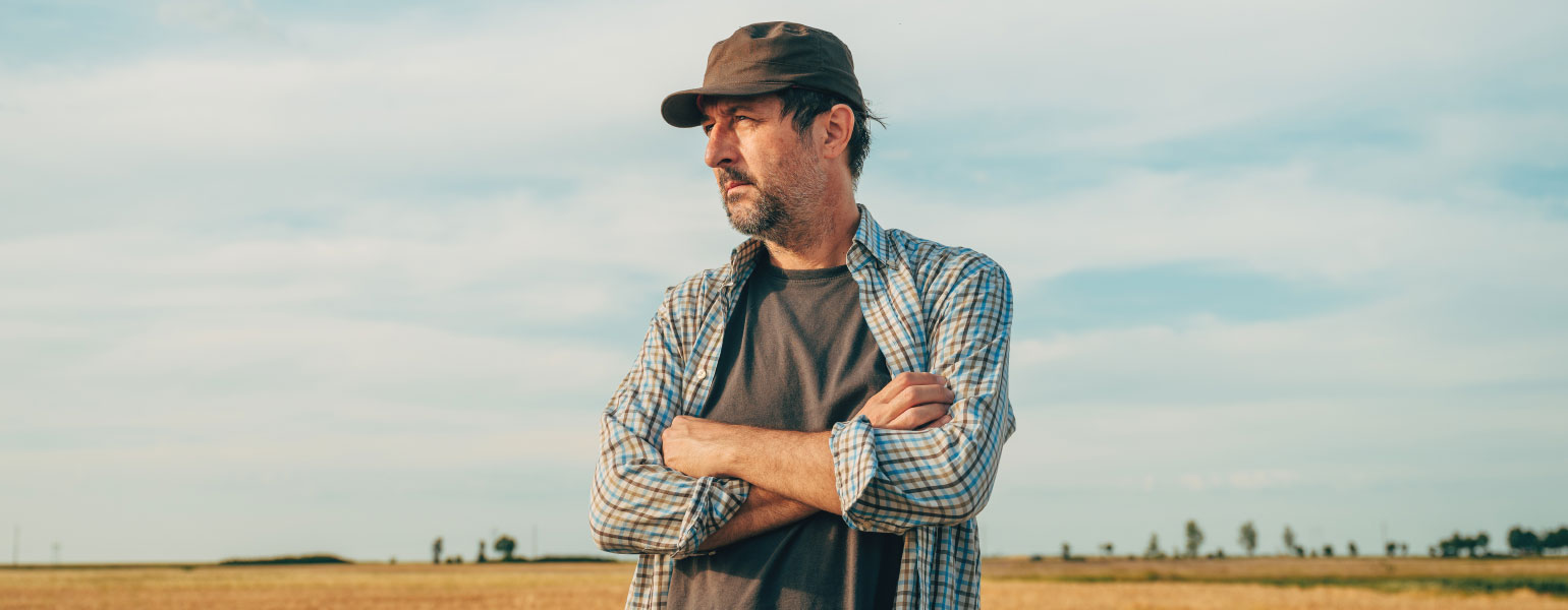 Farmer with trucker's hat standing in ripe wheat crop field with arms crossed